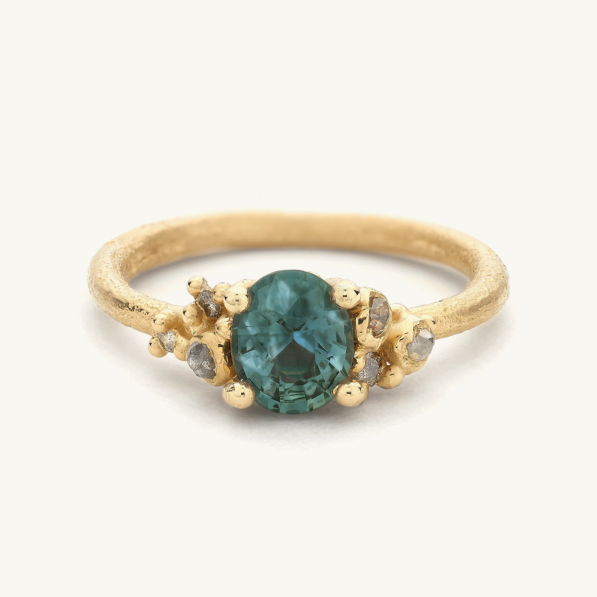 Handcrafted engagement ring by Ruth Tomlinson featuring an oval cut teal-green sapphire framed by clusters of mixed cut diamonds. 
